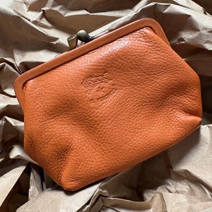May include: A textured orange leather coin purse with a kiss-lock closure featuring a metallic bronze frame. The front of the purse displays an embossed logo of a bison and the text IL BISONTE, centered below the metal clasp mechanism.