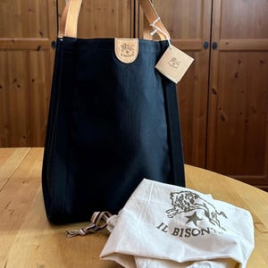 May include: A black tote bag with tan leather handles and a leather patch with a logo. A white drawstring bag with a logo is next to the tote bag. The tote bag has a tag attached.