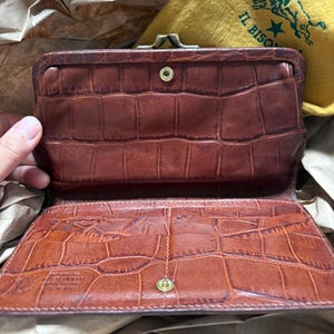 May include: A brown leather wallet with a crocodile-embossed texture, featuring a metal kiss-lock closure at the top and a snap button fastener. The interior displays a stamped logo and the text IL BISONTE on a yellow fabric pouch nearby.