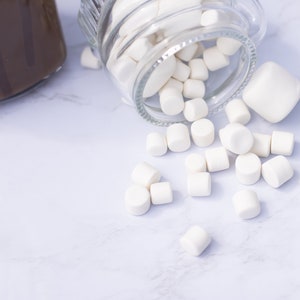 Könnte beinhalten: Ein Glas gefüllt mit weißen Marshmallows kippt auf eine weiße Marmorfläche. Einige Marshmallows sind noch im Glas.