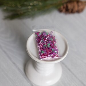 Puede incluir: Una pequeña bolsa de plástico transparente llena de espolvorear rosa y morado. Las chispas tienen forma de pequeñas bayas y palitos largos y delgados. La bolsa está sentada en un soporte para pasteles blanco.