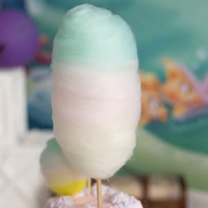 May include: A large, fluffy cotton candy swirl with a light blue top layer, a pink middle layer, and a white bottom layer. The cotton candy is on a stick and is ready to be eaten.