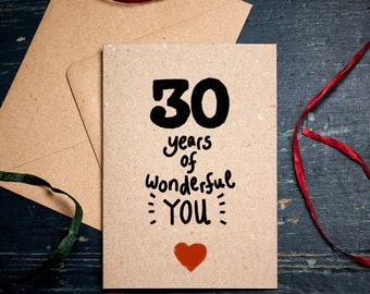 May include: A kraft paper greeting card with the text "30 years of Wonderful YOU" in black script and a red heart. The card is accompanied by a matching envelope and red ribbon on a dark blue wooden surface.