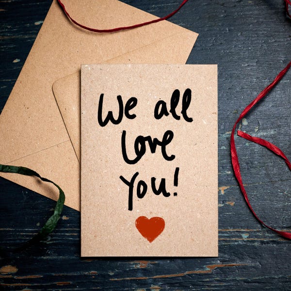 Peut inclure: Une carte brune avec les mots "We all love you!" écrits en noir. Un cœur rouge est dessiné sous le texte.