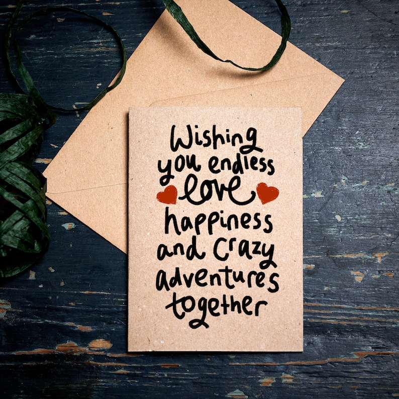 May include: A brown card with the text "Wishing you endless love happiness and crazy adventures together" written in black marker. Two red hearts are drawn on either side of the word "love". The card is sitting on a brown envelope with a green ribbon.