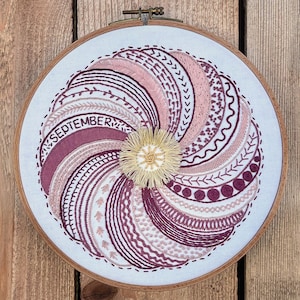 May include: Embroidered hoop art with a swirling design in shades of pink and purple. The design includes the word "SEPTEMBER" and a floral motif in the center.