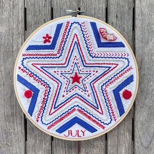 May include: A white embroidery hoop with a red, white, and blue star design. The star is made up of different stitches and the word "JULY" is embroidered in red at the bottom of the hoop.