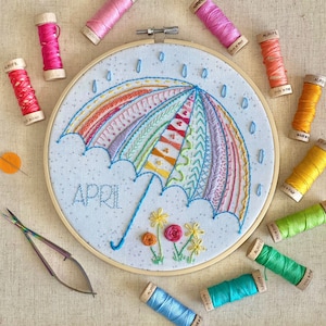 May include: A colorful embroidery hoop with a rainbow umbrella, flowers, and the word "APRIL" stitched on a white fabric background. The hoop is surrounded by spools of embroidery floss in various colors.