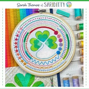 May include: A white embroidery hoop with a colorful embroidery design of a shamrock. The hoop is surrounded by spools of embroidery floss in various colors.