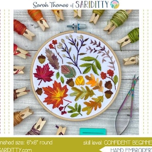 May include: A hand embroidery pattern featuring a 6"x6" round design with autumn leaves and acorns. The design is stitched on white fabric within an embroidery hoop. The image includes embroidery floss, clothespins, and scissors. The text reads "SCATTERED STITCH".