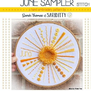 May include: A hand embroidery pattern for June, featuring a sun with yellow and orange embroidery stitches. The pattern is stitched on a white fabric and framed in a wooden hoop. The text "June" is stitched in orange thread below the sun.