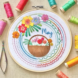 May include: Embroidered hoop art featuring a basket of flowers with the word "May" in the center. The hoop is surrounded by colorful embroidery stitches in a circular pattern.