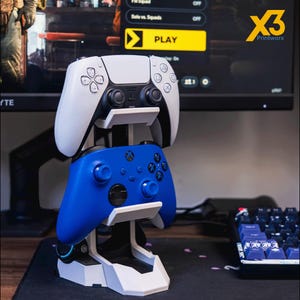 May include: A white and blue gaming controller stand holding a white PlayStation 5 controller and a blue Xbox controller. The stand is white and black, with the text "X3 Printworx" in the upper right corner.