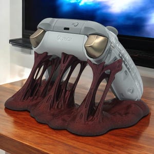 May include: A gray Xbox controller rests on a maroon-colored stand. The controller has gold accents and the Xbox logo. The stand has a unique, textured design with a flowing, organic shape, creating a striking contrast.