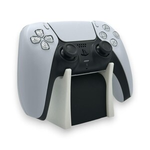 以下が含まれることがあります： 白と黒のPlayStation 5コントローラーが白いスタンドに置かれています。コントローラーには、黒いジョイスティックとボタン、方向パッド、アクションボタンがあります。スタンドには黒いベースがあります。