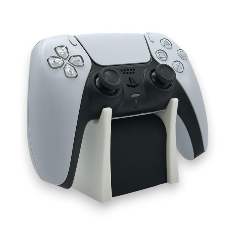 以下が含まれることがあります： 白と黒のPlayStation 5コントローラーが白いスタンドに置かれています。コントローラーには、黒いジョイスティックとボタン、方向パッド、アクションボタンがあります。スタンドには黒いベースがあります。
