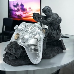 May include: A clear Xbox controller with gold accents and a black soldier statue. The controller sits on a black, textured base. The soldier is kneeling and holding a gun. A television is visible in the background.