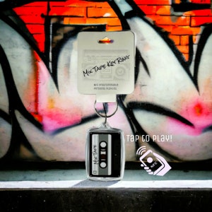 May include: A clear acrylic Mix Tape Key Ring with a black and gray cassette tape design. The key ring includes the text "MIX TAPE" and "TAP TO PLAY!" with an NFC symbol. The background features graffiti art.