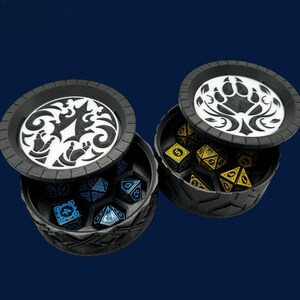 May include: Two black dice boxes with decorative lids, each containing polyhedral dice. One box holds blue dice with white numbers, the other holds yellow dice with black numbers. The lids feature white and black graphic designs.