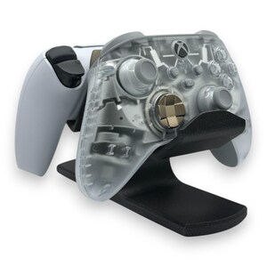 May include: A clear Xbox controller with gold accents rests on a black stand, next to a white controller. The transparent controller reveals internal components. The stand is made of a matte black material.