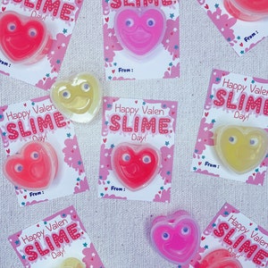 May include: Nine Valentine's Day slime favors with heart-shaped containers. Each container has googly eyes and a smiling face. The containers are pink, red, and yellow. The cards have a pink and white background with stars and clouds. The cards say "Happy Valentine's Day Slime!"