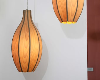May include: Two pendant lights with a wooden veneer design. The lights have a teardrop shape with vertical dark brown stripes. The interior glows with a warm light. The lights hang from thin silver cords against a white wall.
