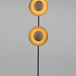 May include: A modern floor lamp with a black metal frame. It features two circular light fixtures with a warm, golden glow, each with a central, darker disc. The lamp stands on a round base.