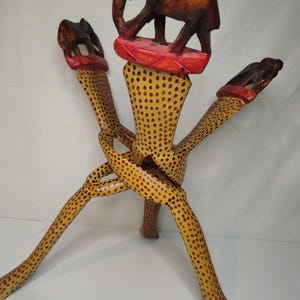 May include: A handcrafted wooden decorative stand featuring an elephant carving at the top. The stand has three legs with a spotted pattern and is adorned with red accents. The elephant carving is brown, and the stand is approximately 12 inches tall.