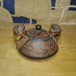 May include: A round, lidded ceramic vessel featuring two sculpted animal head handles on opposite sides. The exterior is decorated with intricate black, red, and tan geometric patterns. A small, dark brown knob sits atop the circular lid.