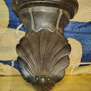 May include: A dark brown wall-mounted shelf featuring a semi-circular top platform and a decorative support bracket carved in the shape of a large, prominent scallop shell with vertical ridges and a detailed base ornament.