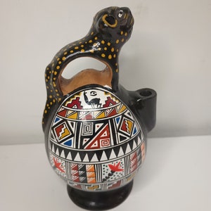 May include: A black ceramic pitcher with a geometric pattern in red, white, blue, yellow, and orange. The pitcher has a handle shaped like a black and yellow spotted animal.