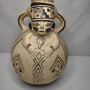 May include: A beige ceramic vessel with a human-like face and geometric patterns. The vessel has a bulbous body, two handles, and a detailed headdress. The design includes black and red accents, creating a unique decorative piece.