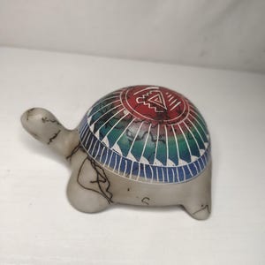 May include: A small, decorative turtle figurine with a painted shell. The shell features a geometric design in red, green, and blue. The turtle is light gray with dark lines.