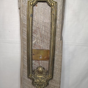 May include: An antique brass door plate with ornate detailing. The rectangular plate features a decorative frame with a beaded border and floral accents at the top and bottom. The plate is mounted on a weathered, light-colored wooden surface.