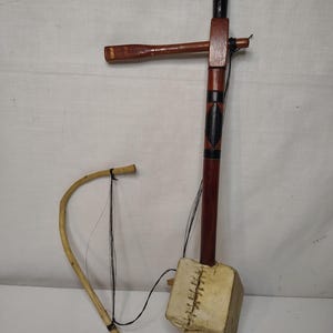 May include: A stringed musical instrument featuring a long, dark brown wooden neck with black accents, a square-shaped resonator covered in light-colored, stitched hide, and a separate curved wooden bow with thin black strings attached to the instrument.