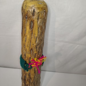 May include: A tall, cylindrical wooden object with a textured, light brown surface. A green knitted band with pink and yellow yarn accents is wrapped around the middle. The top is a lighter color.