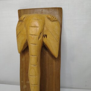 May include: A wooden elephant sculpture with a light brown finish. The elephant is carved in a detailed, three-dimensional style and mounted on a rectangular wooden plaque. The elephant's trunk is extended downward.