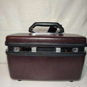 May include: A burgundy vintage suitcase with a black handle and silver-toned accents. The rectangular case has a textured surface and two silver latches. The handle is attached with silver metal and a black strap.