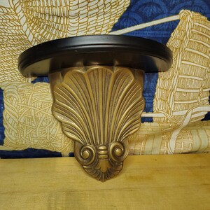 May include: A wall-mounted shelf featuring a smooth, semi-circular black top surface supported by a decorative, gold-toned bracket. The bracket is molded into a prominent, symmetrical seashell design with vertical ridges and scrolled base details.
