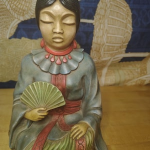 May include: A ceramic figurine of a person with dark hair in a bun, wearing a blue robe with a red collar and belt, and a green skirt. The figure holds a small, light green handheld fan and has closed eyes, featuring detailed painted accents.