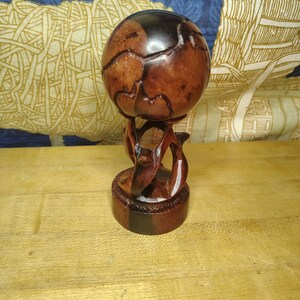 May include: A dark brown wooden sphere with natural grain patterns and deep fissures, resting on a carved wooden stand featuring three curved, interlocking supports that rise from a circular base with a decorative notched edge.