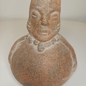 May include: A terracotta figurine of a person with a round body and a large head. The figurine has a simple, stylized face with closed eyes and a small mouth. The figurine is decorated with a beaded necklace around the neck.