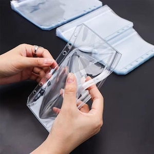 May include: Clear plastic binder pockets with a zip closure. The pockets have reinforced holes for easy insertion into a binder. The image shows a person opening the zip of one pocket, with several other pockets in the background.