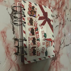 Puede incluir: Un cuaderno con espiral con temática navideña. La portada presenta un lazo rojo, vagones de tren de pan de jengibre y el texto "25 Days of Christmas". El cuaderno tiene un fondo blanco con rayas de borde rojas y grises.