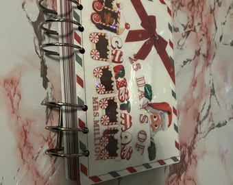 May include: A spiral-bound notebook with a Christmas theme. The cover features a red bow, gingerbread train cars, and the text "25 Days of Christmas". The notebook has a white background with red and gray border stripes.