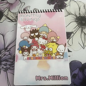 May include: A spiral-bound notebook with a pink cover featuring Sanrio characters. The cover has the text "MONTHLY SAVINGS" at the top and "SANRIO CHARACTERS" at the bottom. The name "Mrs. Million" is printed at the bottom.