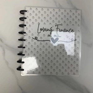 May include: A planner with a clear cover and black spiral binding. The cover features the words "Loving Finance" and "Mr. Millien" in script, along with a heart graphic. The background has a pattern of small, repeating images.
