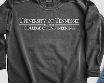 May include: A dark grey sweatshirt featuring the white text "UNIVERSITY OF TENNESSEE EST. 1838 COLLEGE OF ENGINEERING". The sweatshirt is shown with blue jeans and white trainers.