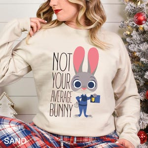 Op de afbeelding: Zandkleurige sweatshirt met de tekst "NOT YOUR AVERAGE BUNNY" en een cartoon konijn. Het konijn heeft roze oren, blauwe ogen, draagt een blauw uniform en houdt een blauwe aktetas vast.