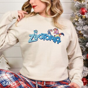 May include: A sand-coloured sweatshirt with the word "Zootopia" in blue, and cartoon characters. The sweatshirt has long sleeves and a crew neck. The bottom of the sweatshirt is partially visible, and the word "SAND" is printed on the bottom left.